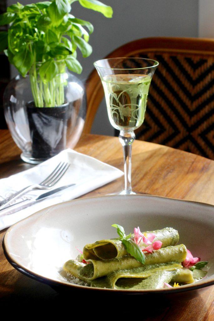 A rustic wooden table set with a bowl of pesto, a plate of gluten-free pasta, and a glass of wine.