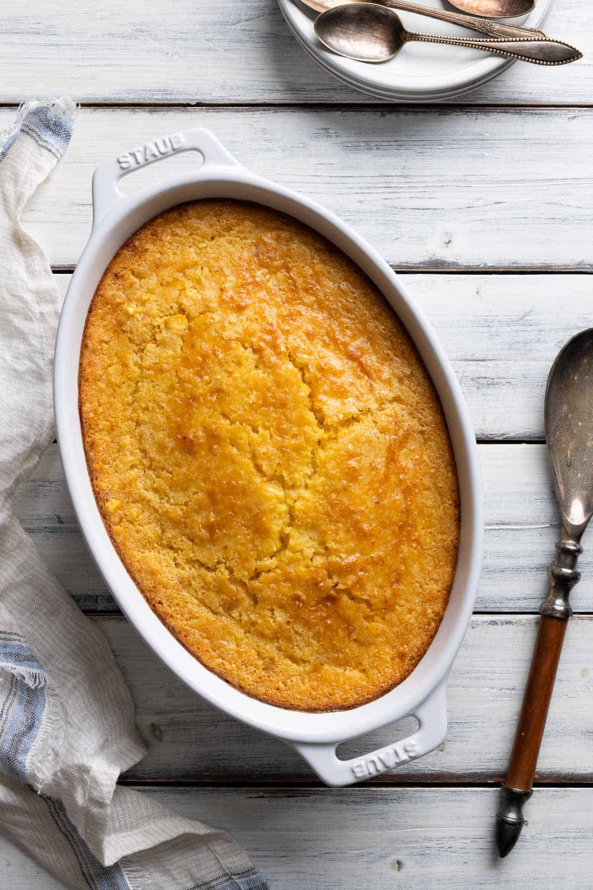 Golden brown corn casserole dish on a rustic wooden table with a serving spoon, holiday setting