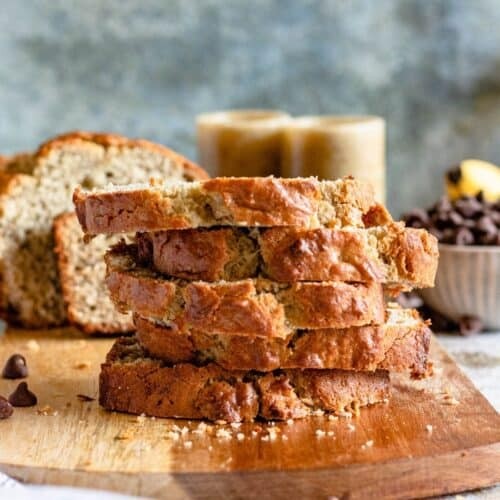 a slice of dark, moist banana bread with visible banana chunks and a golden crust, set against a rustic wooden background with scattered banana peels and a single banana