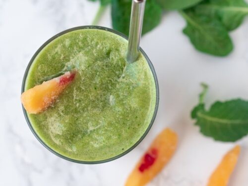 a vibrant green smoothie with peach slices and leafy greens, garnished with mint leaves, in a clear glass on a bright, sunny countertop