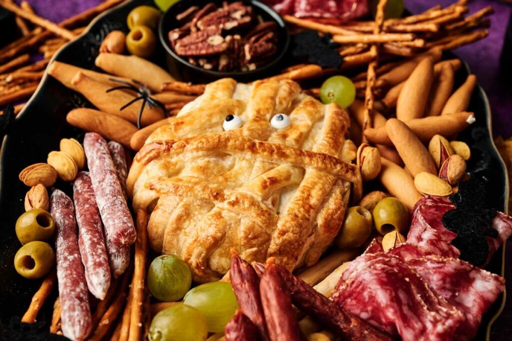 Baked camembert cheese wheel surrounded by charcuterie meats, crackers, fruit, and nuts on a wooden board