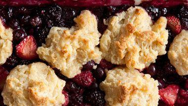 A rustic baking dish filled with a golden-brown berry crumble topped with a dusting of powdered sugar, garnished with fresh lingonberries.