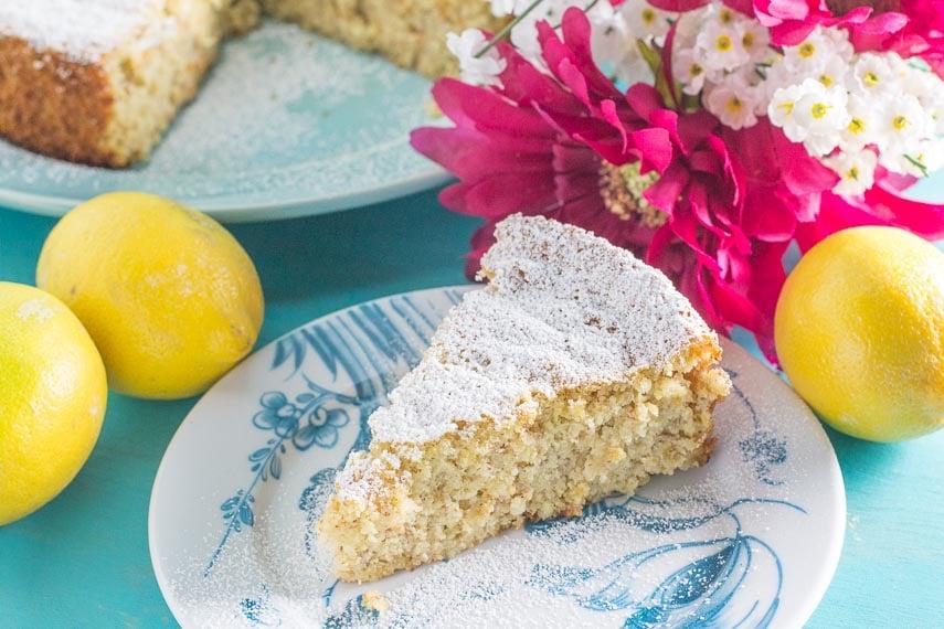 A slice of lemon almond cake on a plate, with a cup of tea in the background.