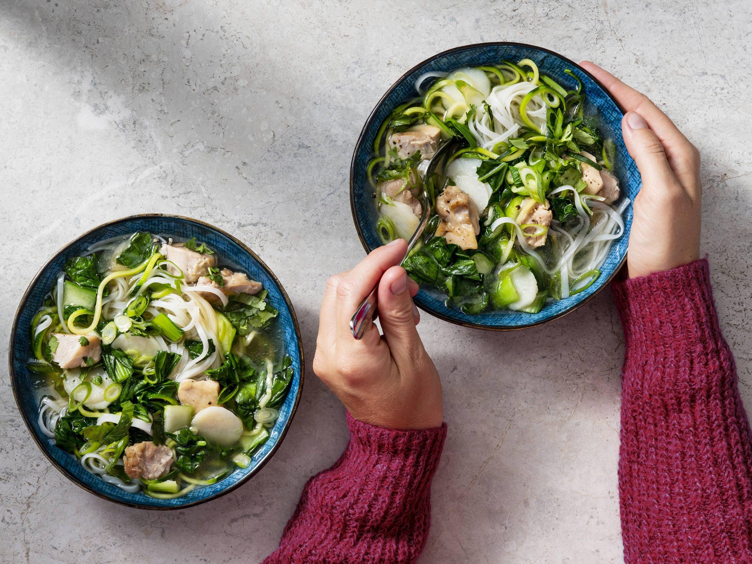 a person enjoying a bowl of steamed rice noodle soup with vegetables and chicken